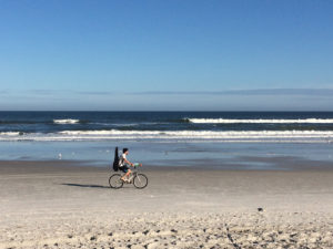 a man rides his bike with a guitar on his back on the sand at atlantic beach in florida, photographed by jamie bannon photography.