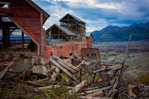 a dilapidated building falls in front of a beautiful mountain landscape in kennecott, alaska, photographed by jamie bannon photography.