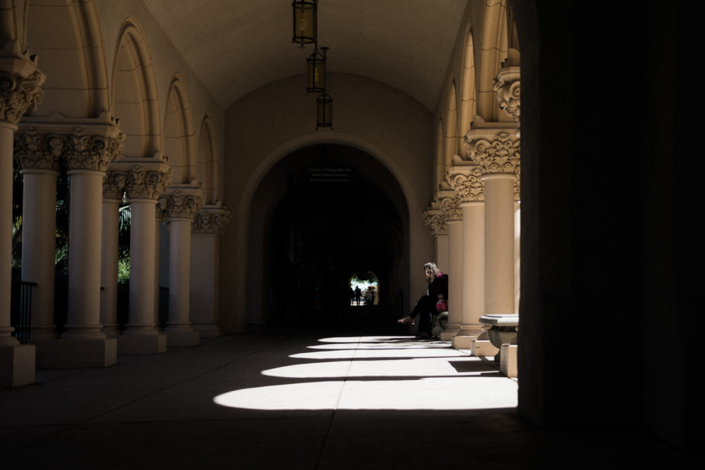 a woman sits amongst the shadows of a decorative colonnade in balboa park, san diego, california, photographed by jamie bannon photography.