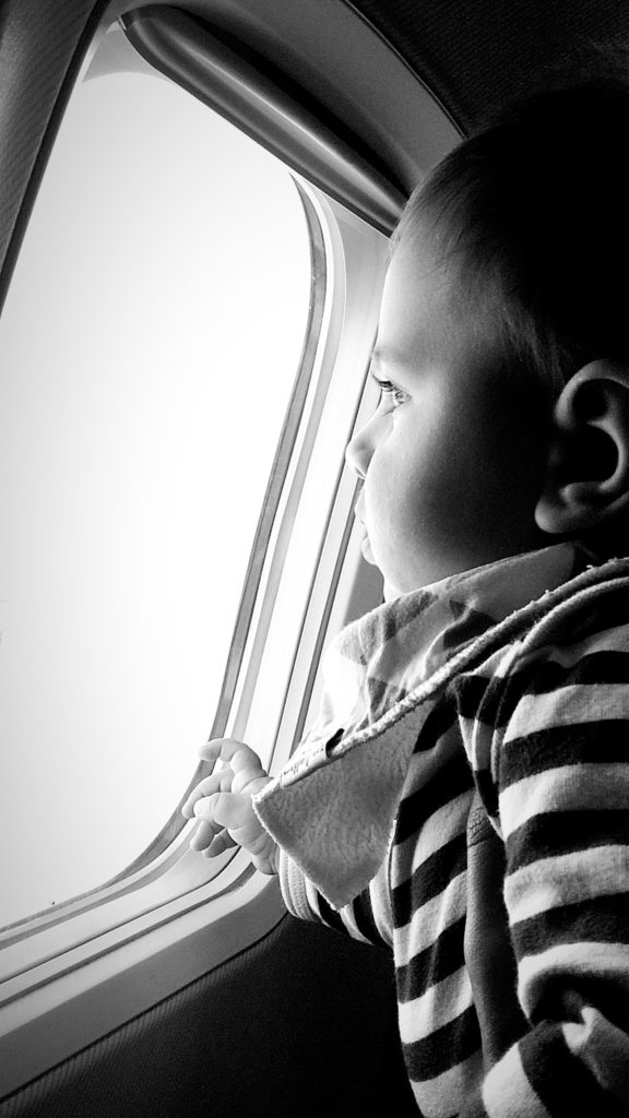black and white portrait of a baby looking out an airplane window on his first flight, photographed by jamie bannon photography.
