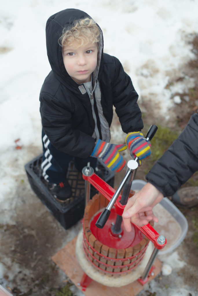 a young boy turns an apple cider press with all his might on a cold new england winter day, photographed by jamie bannon photography.